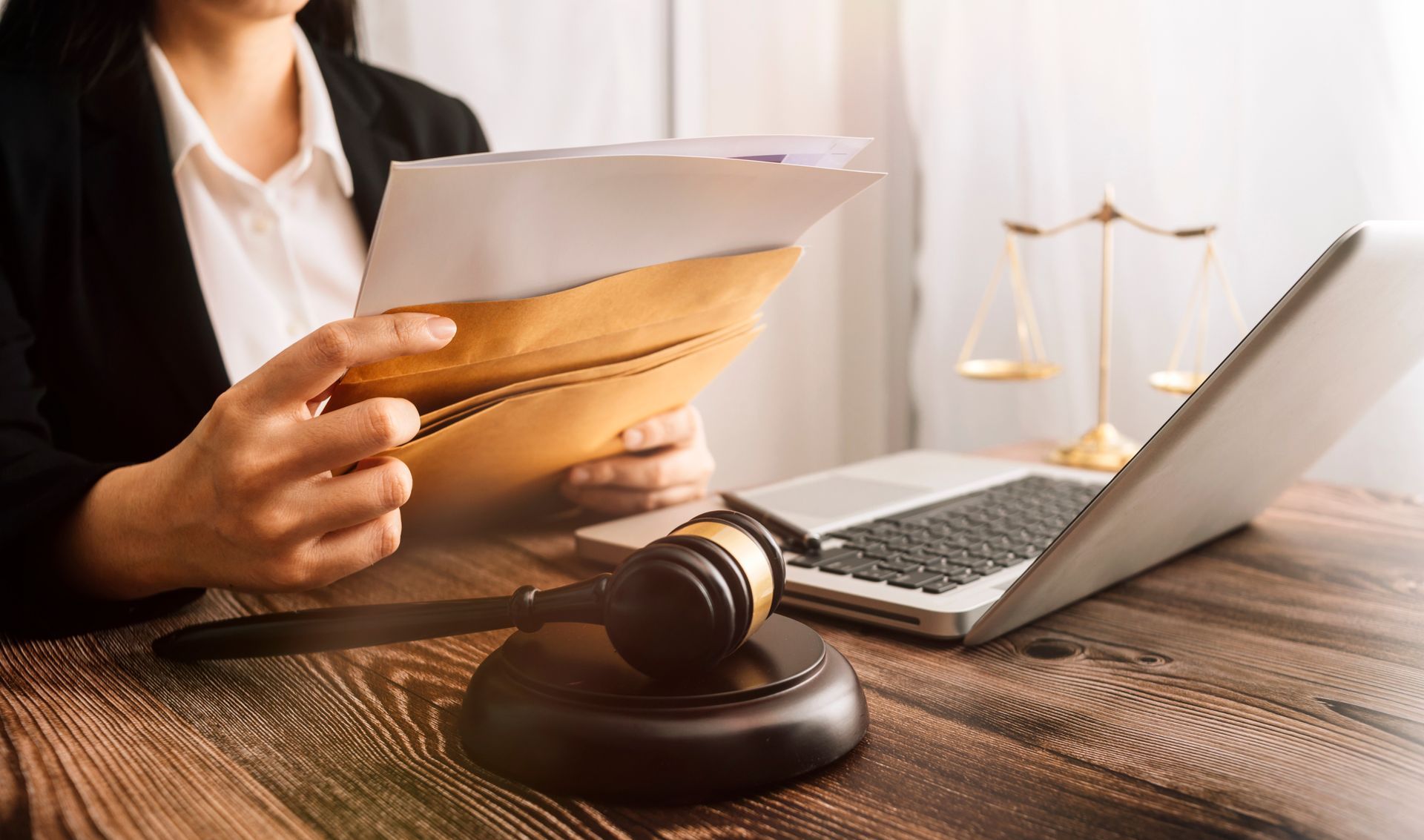 Person in a suit holding documents beside a laptop and judge’s gavel on a desk Person in a suit holding documents beside a laptop and judge’s gavel on a desk