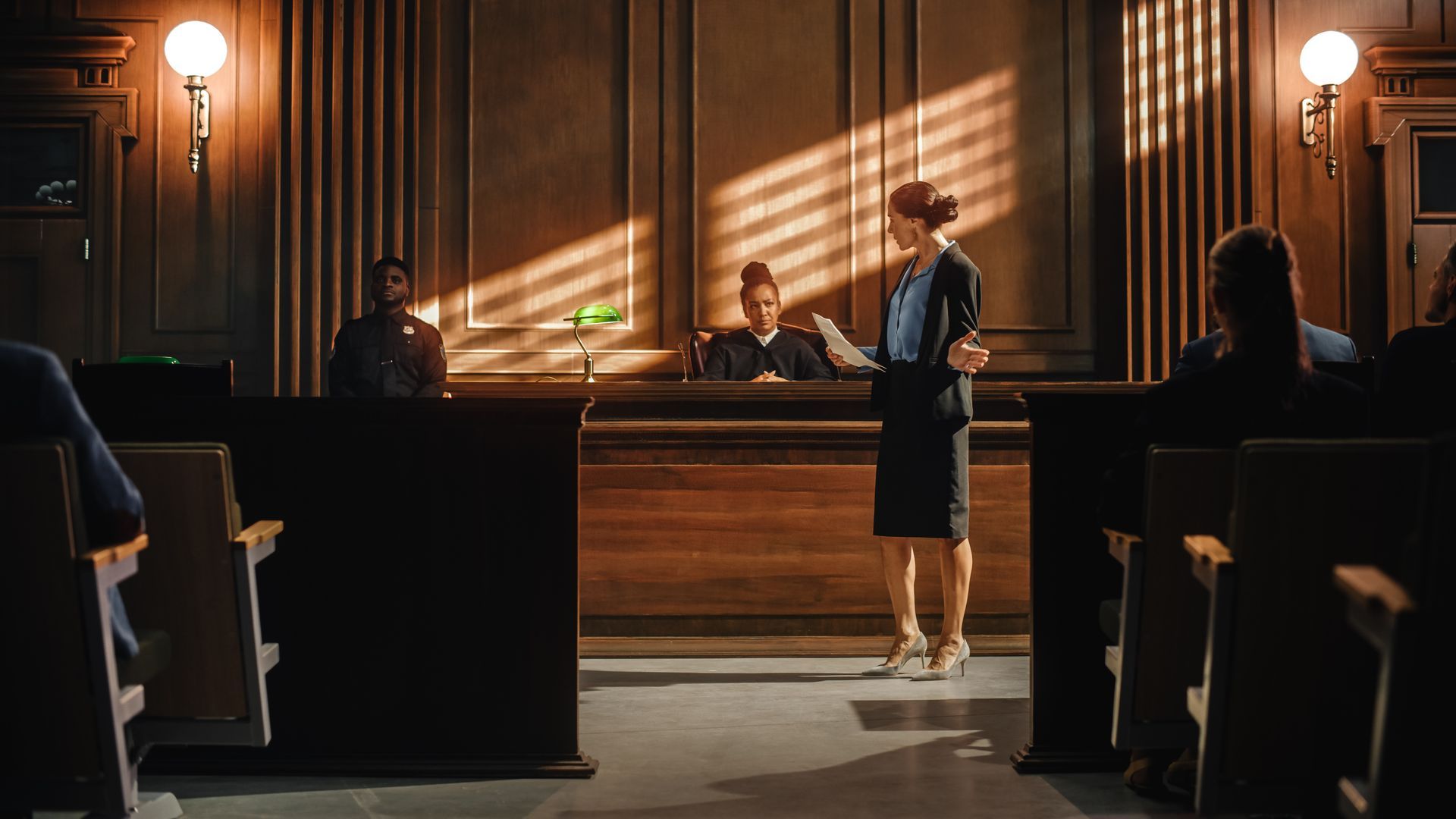 Courtroom with judge at bench, lawyer standing before it, and seated attendees in dark wood interior. Courtroom with judge at bench, lawyer standing before it, and seated attendees in dark wood interior.