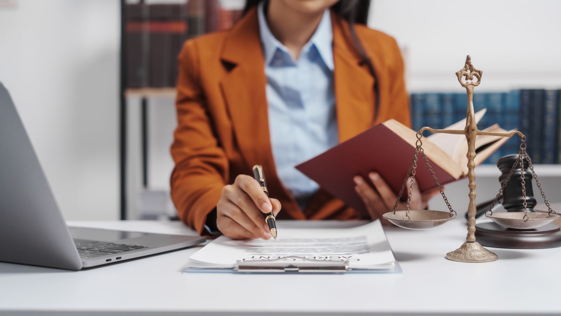 Legal professional writing at a desk with laptop, notebook, scales of justice, and law book Legal professional writing at a desk with laptop, notebook, scales of justice, and law book