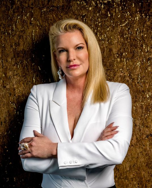 Woman in a white blazer, arms crossed, posing against a textured gold backdrop.