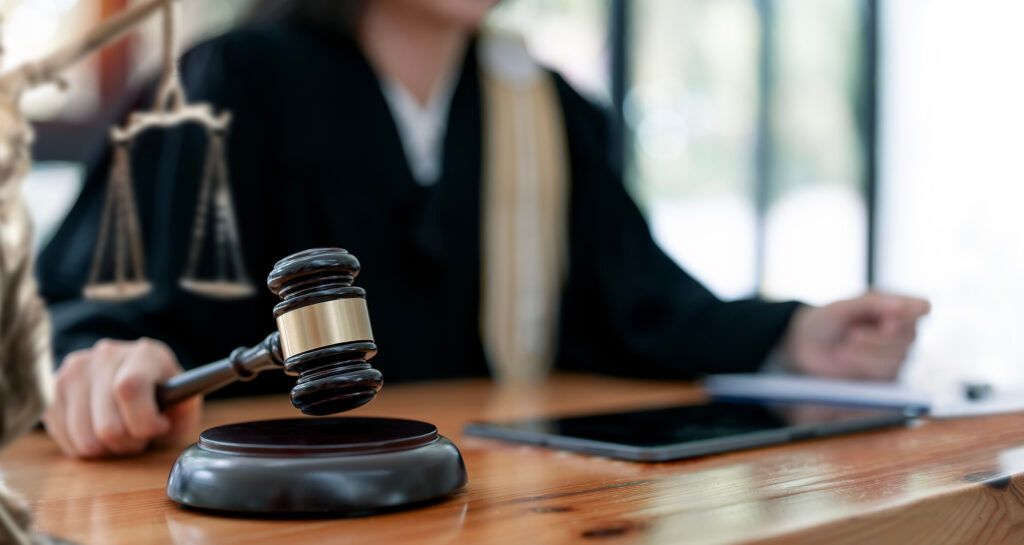 Judgeโs gavel on a wooden desk with a blurred lawyer in the background and a tablet nearby