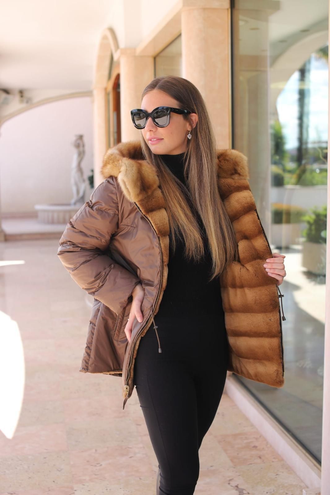 Donna con occhiali da sole e piumino in posa all'aperto accanto a una parete di vetro.