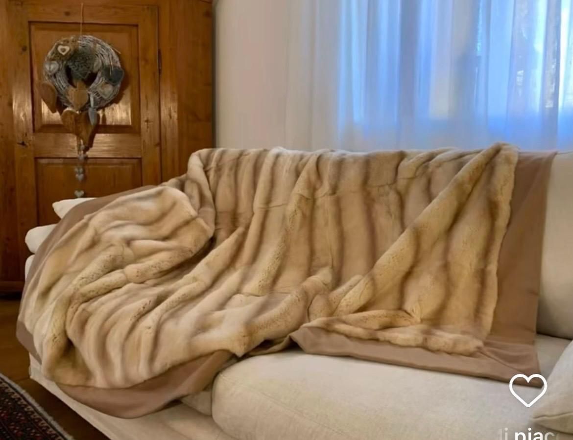 Un divano beige drappeggiato con una coperta di pelliccia sintetica marrone accanto a una porta di legno e tende bianche.