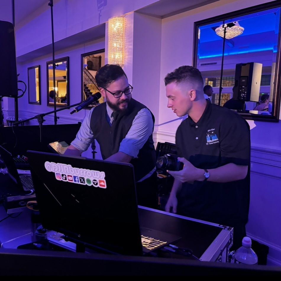 Two professionals work together at a DJ booth in a dimly lit, purple-hued event hall.