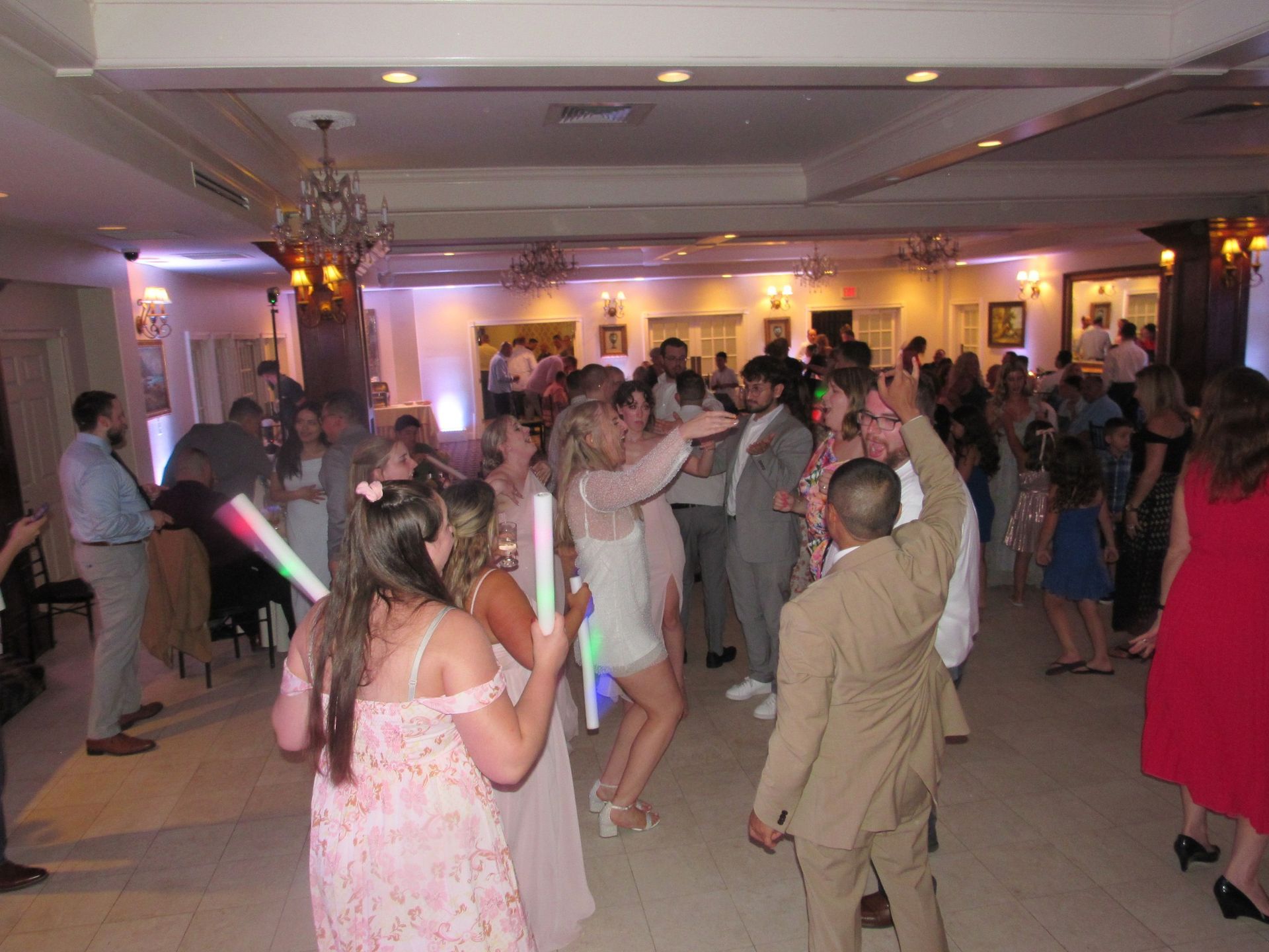 Guests dance in a ballroom, with some holding glow sticks under warm interior lighting.