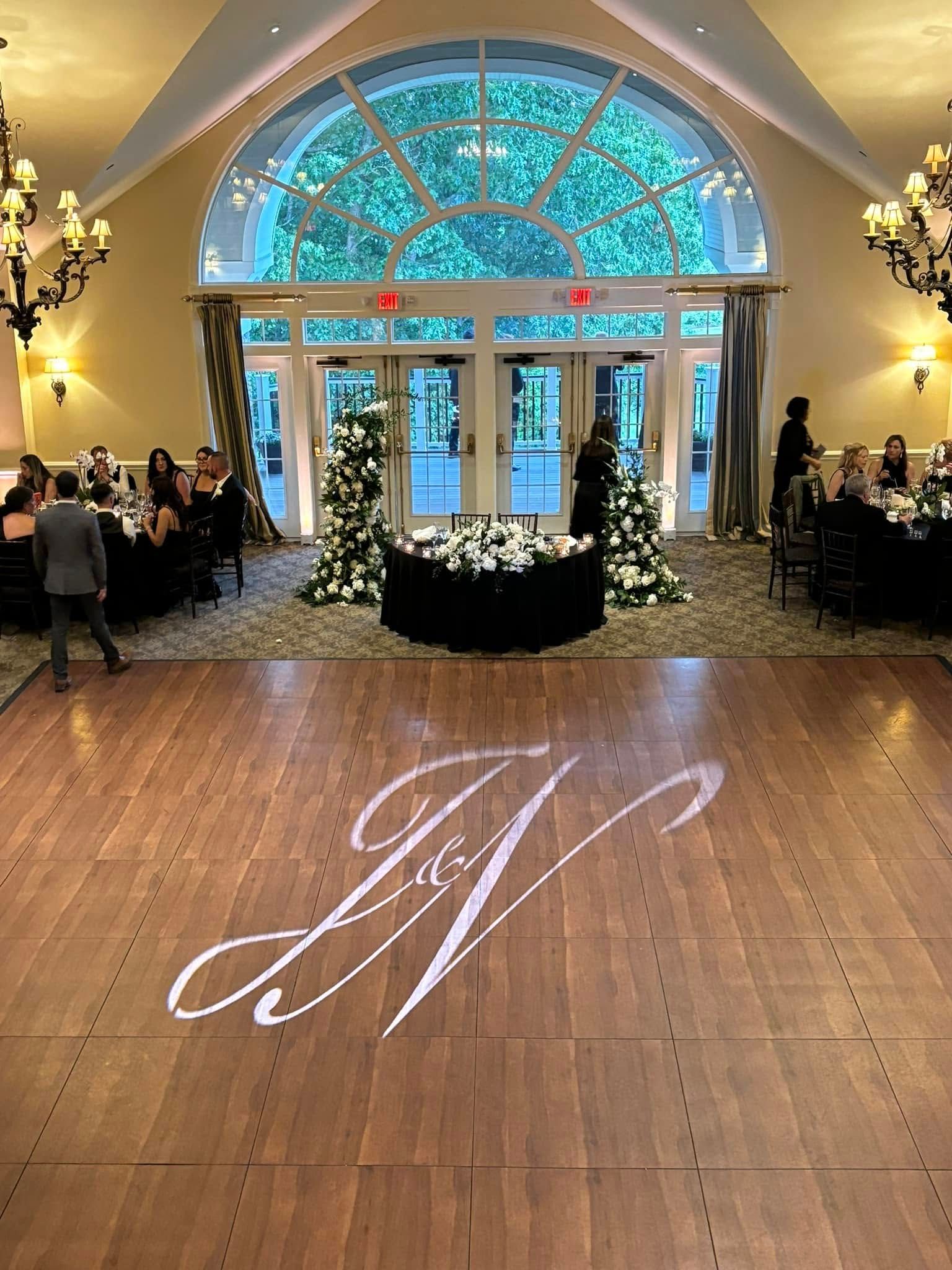 A bright wedding reception ballroom with a wooden dance floor featuring a monogram light projection and floral decor.