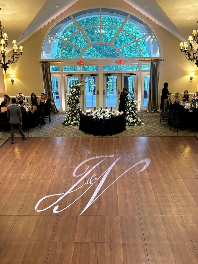 A bright wedding reception ballroom with a wooden dance floor featuring a monogram light projection and floral decor.