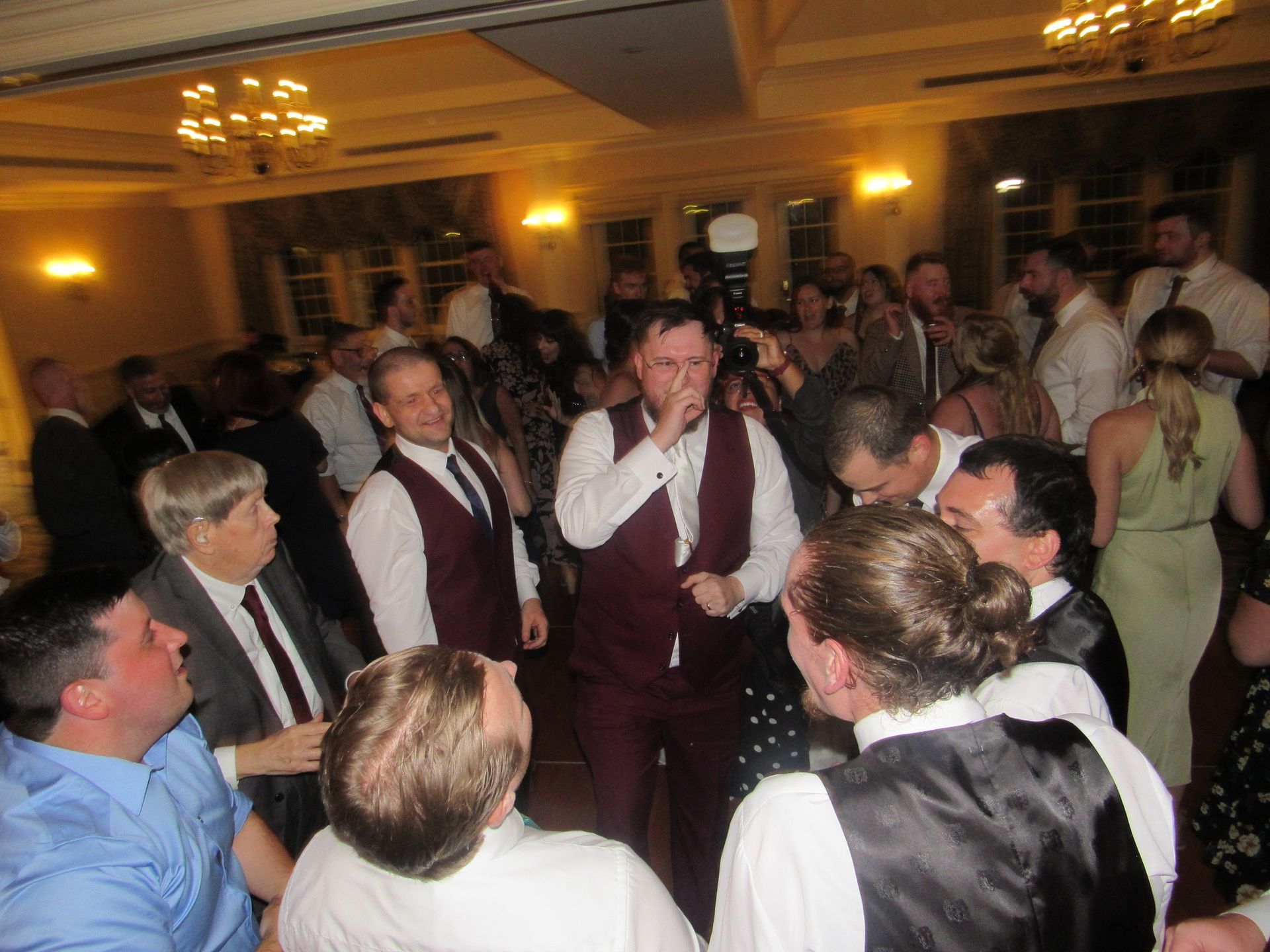 A group of people in formal attire dancing and socializing in a dimly lit ballroom with chandeliers.