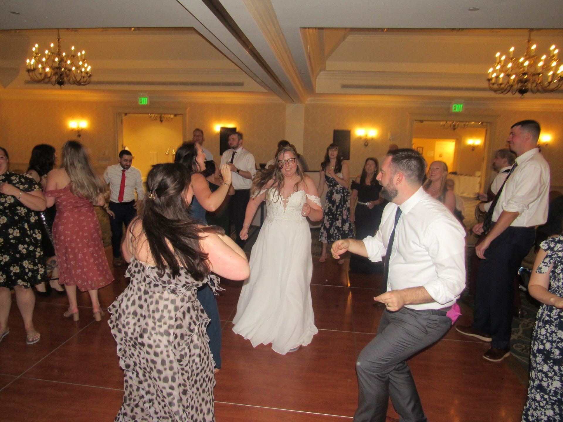 Wedding guests dancing on a wooden dance floor in a ballroom setting, with a bride in a white dress in the center.