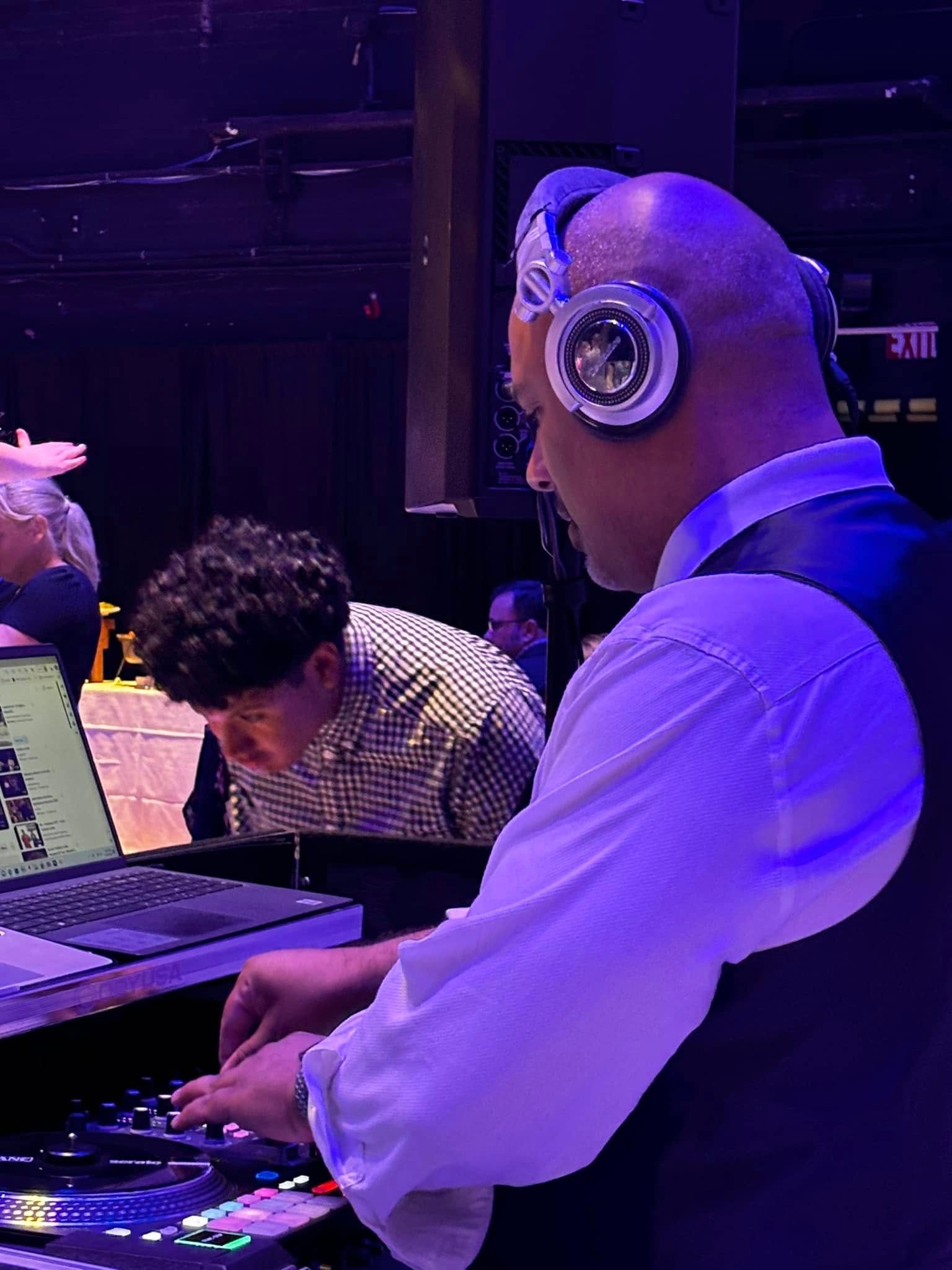 A DJ wearing headphones interacts with a mixing console at a performance, with a person standing at a laptop nearby.