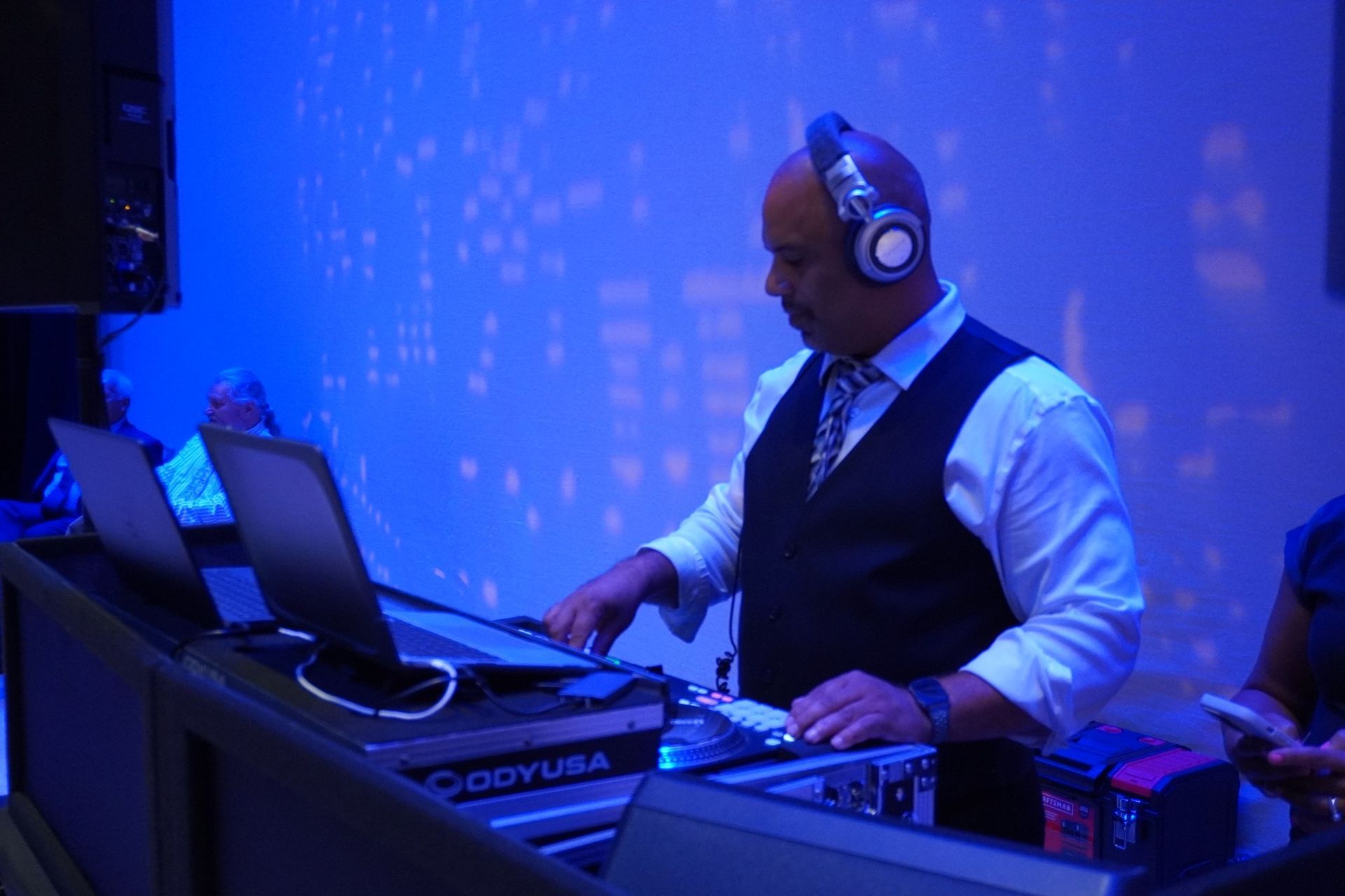 A DJ in a white shirt and vest wearing headphones, operating a laptop and sound equipment in a room with blue lighting.