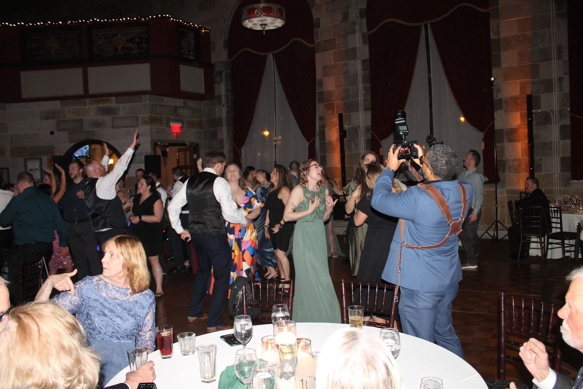 People dance and celebrate at an evening wedding reception inside a hall with stone walls and high windows.