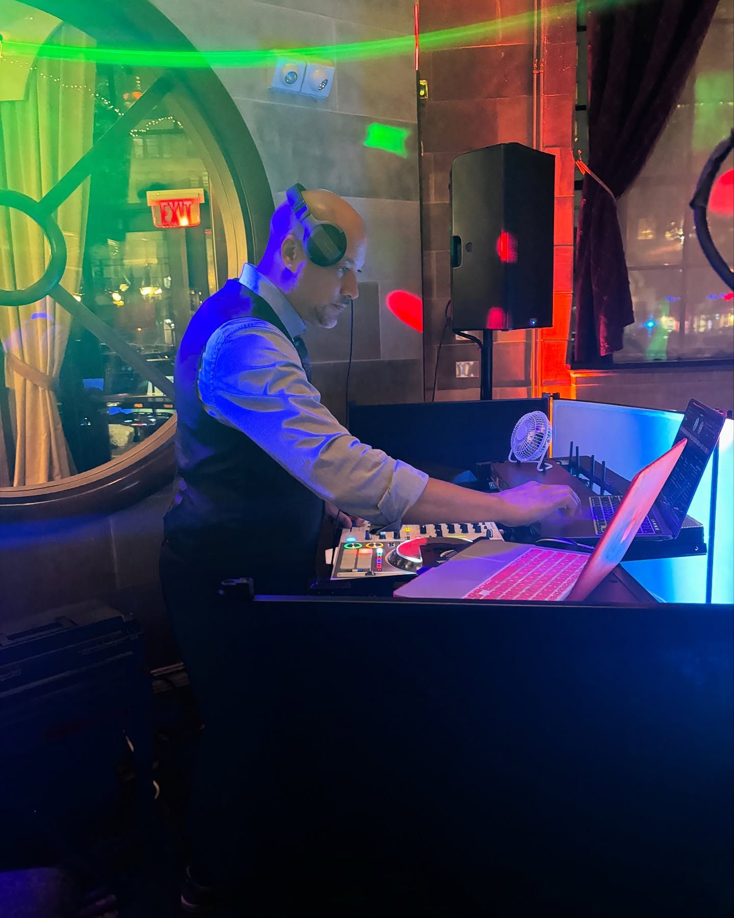 A person wearing headphones and a vest operates DJ equipment and a laptop in a dimly lit venue with colorful lighting.