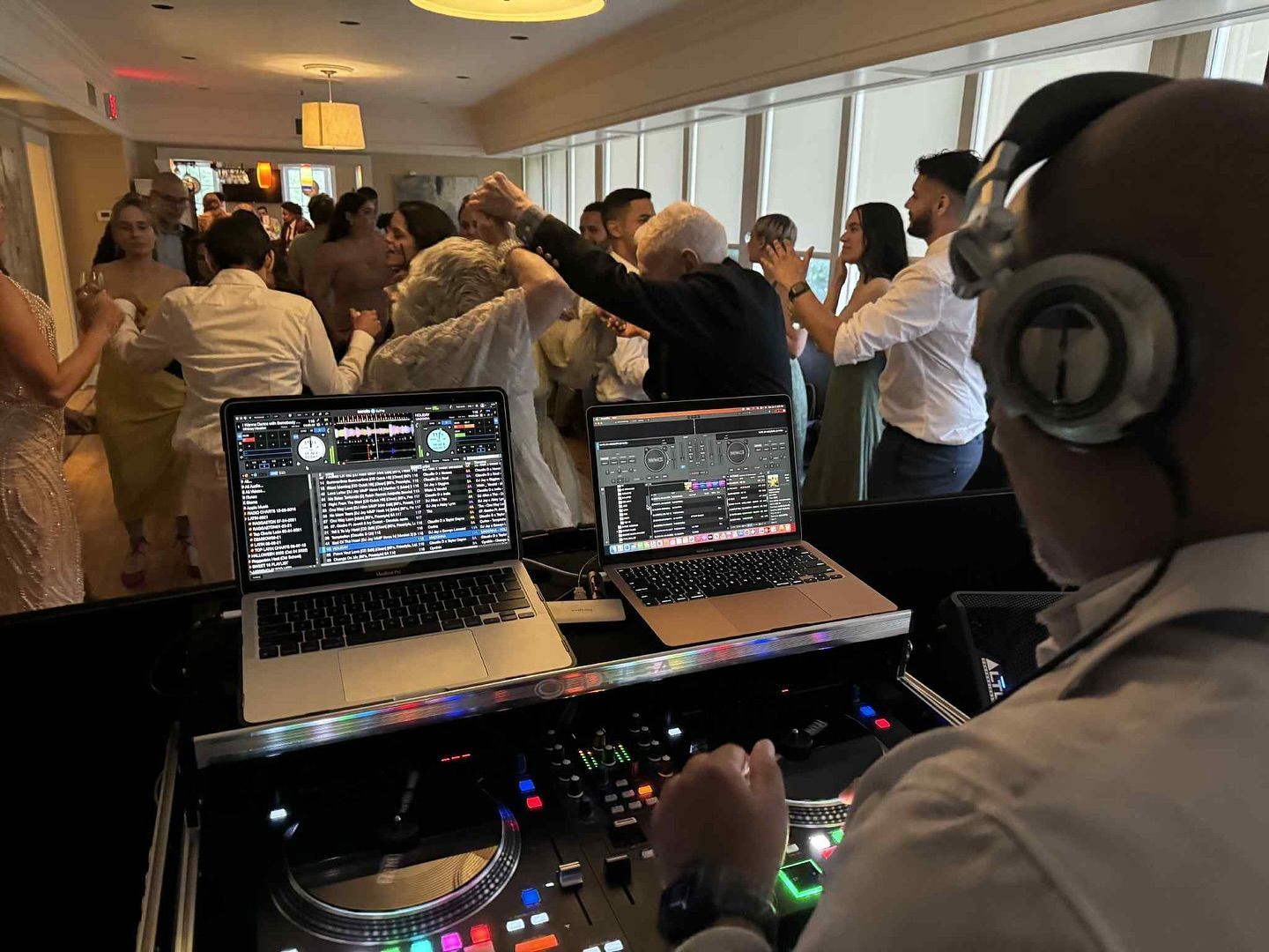 A DJ wearing headphones overlooks a crowded room, mixing music on two laptops and a digital controller for party guests.