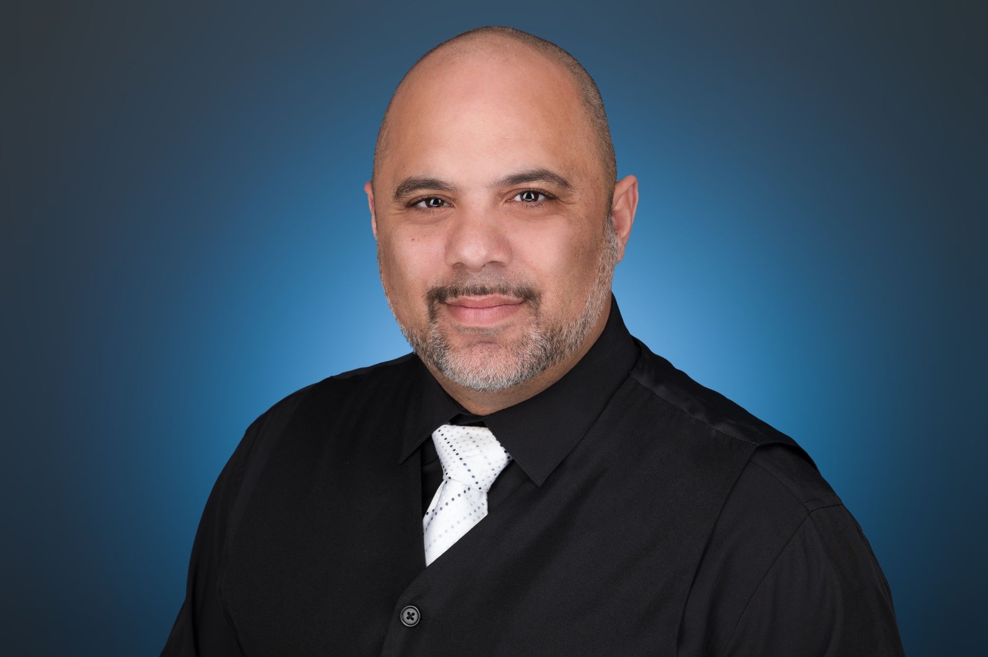 A man with a shaved head and facial hair, wearing a black collared shirt and white patterned tie against a blue background.