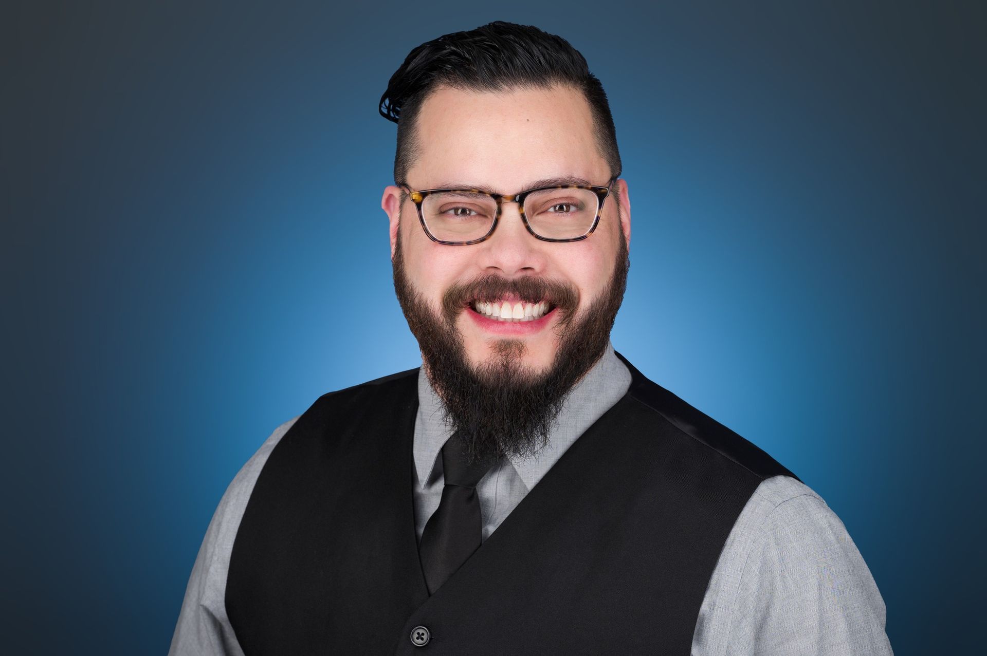 A smiling person with a beard and glasses wears a gray shirt and black vest against a blue gradient background.