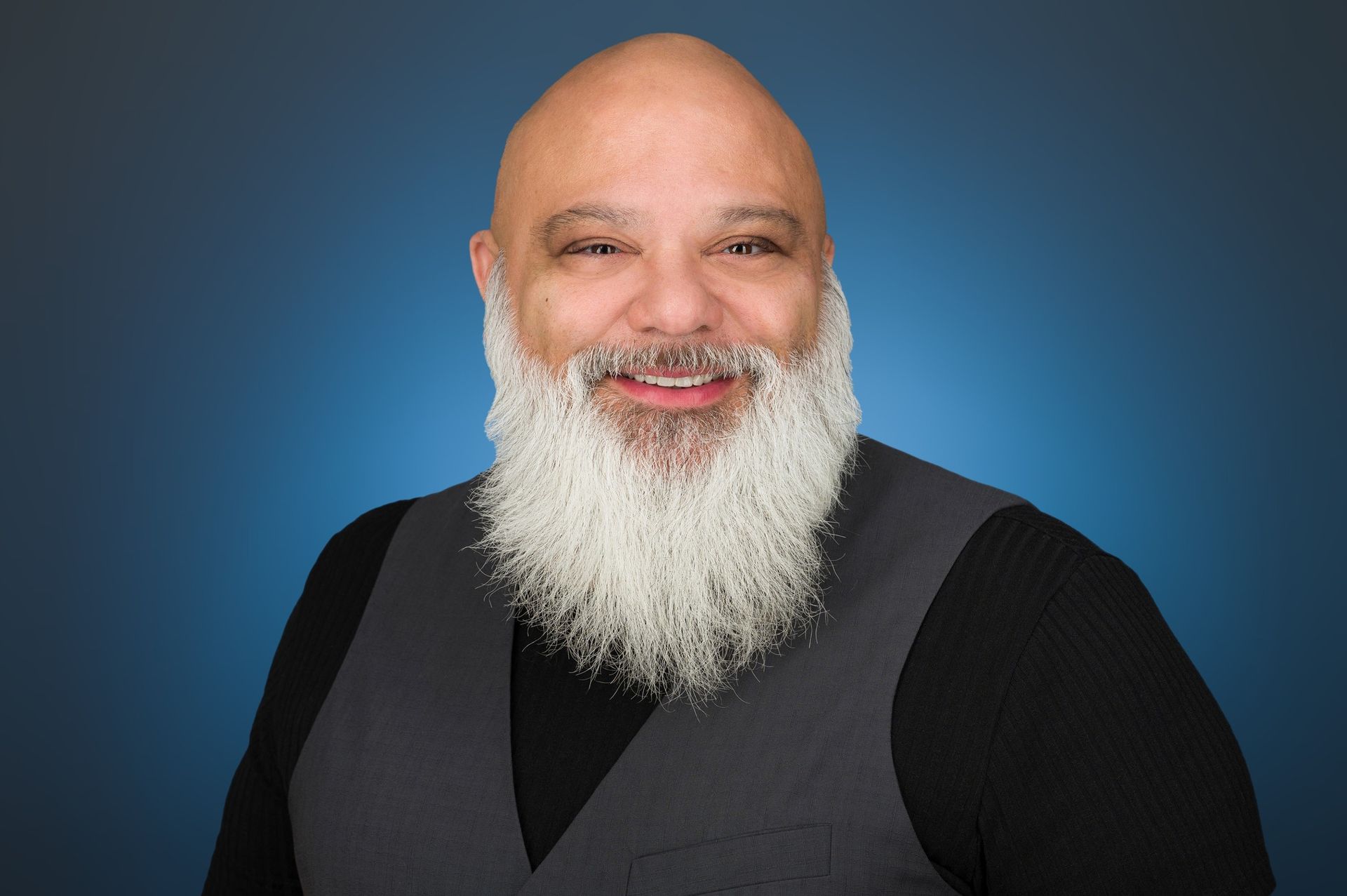 A smiling person with a long white beard, wearing a black shirt and grey vest, against a blue background.