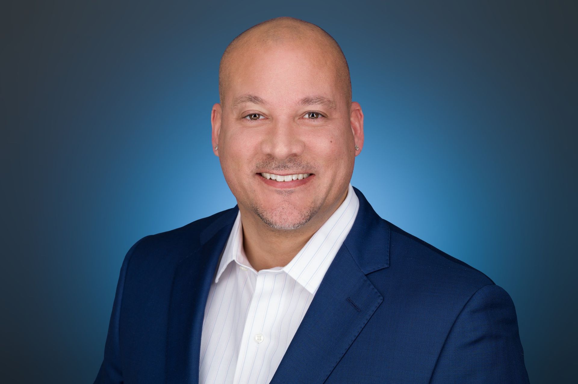 A smiling man with a shaved head wearing a dark blue suit jacket and white dress shirt against a blue gradient background.