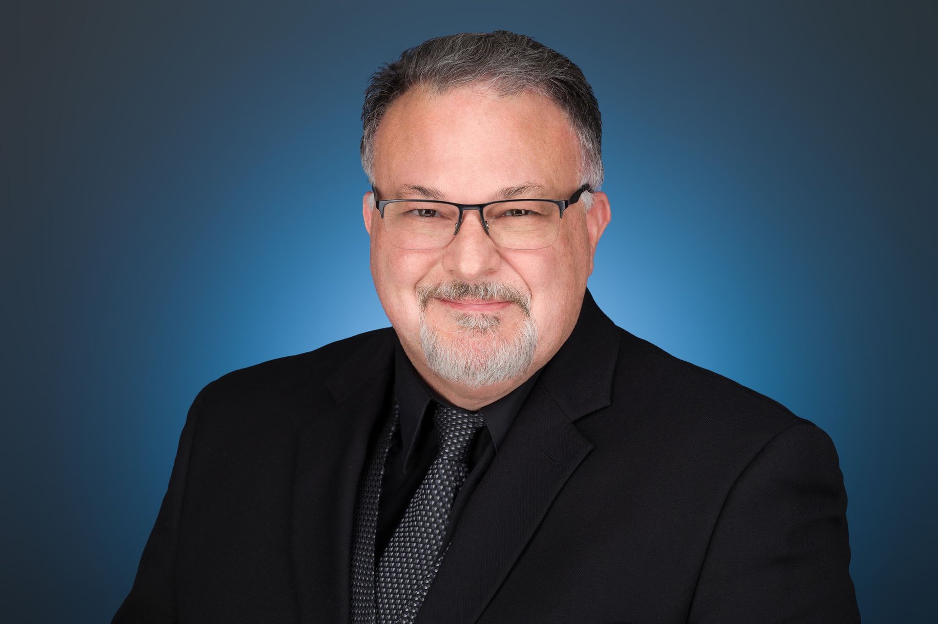 A person wearing a black suit, patterned tie, and glasses, smiling against a blue gradient background.