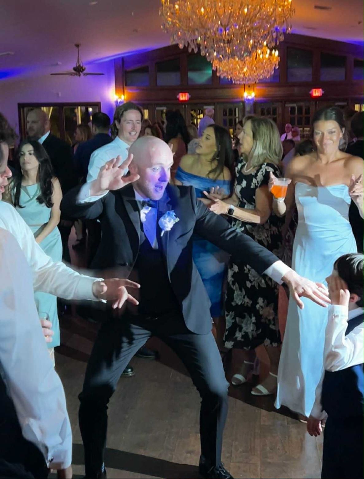 A groom in a black suit dances energetically on a ballroom dance floor among wedding guests under a crystal chandelier.