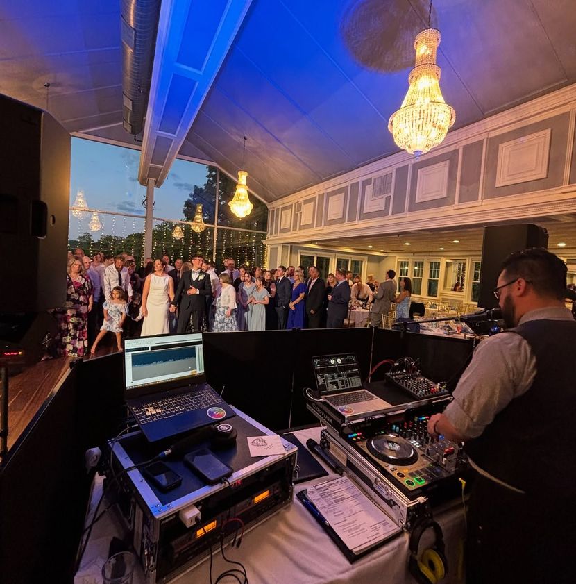A DJ in a vest performs at a wedding reception from behind a mixing console, facing a crowd on a decorated dance floor.