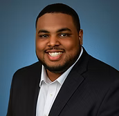 A headshot of a person with a short beard, wearing a dark suit and white dress shirt, smiling against a blue background.