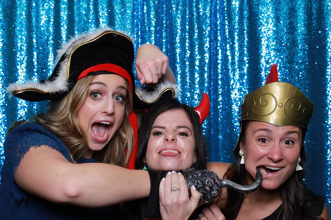 Three women wearing fun costume hats pose playfully in front of a shimmery blue sequin backdrop.