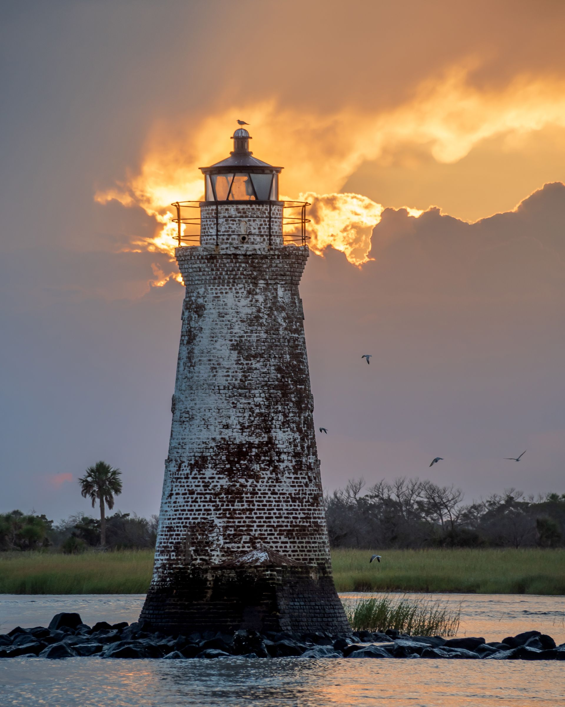 a lighthouse in the middle of a body of water with a sunset in the background . Cockspur Island Lighthouse, Talking Excitingly, Brad Burgess Photography