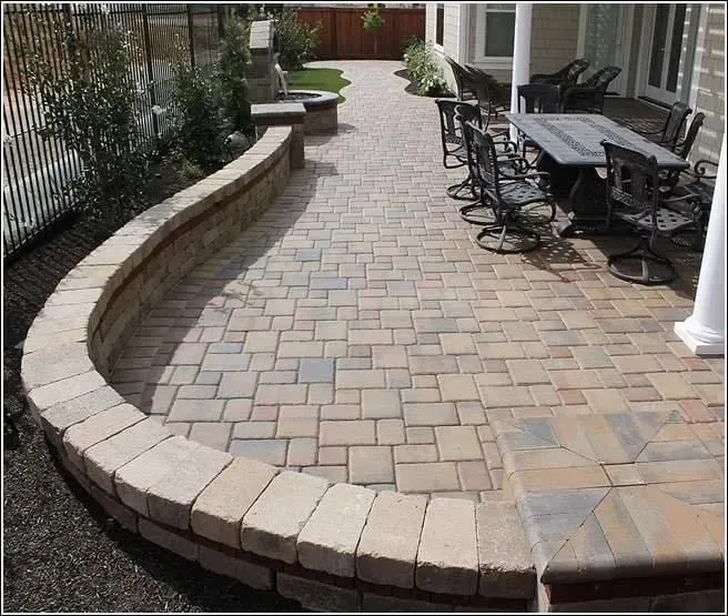 Curved brick-paver patio walkway beside a house, with outdoor table and chairs along the right side.