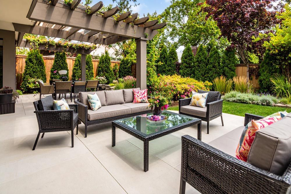 Patio with gray furniture under a wooden pergola, surrounded by lush green garden.