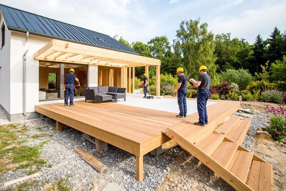 Construction workers on a wooden deck, building near a house with a pergola.