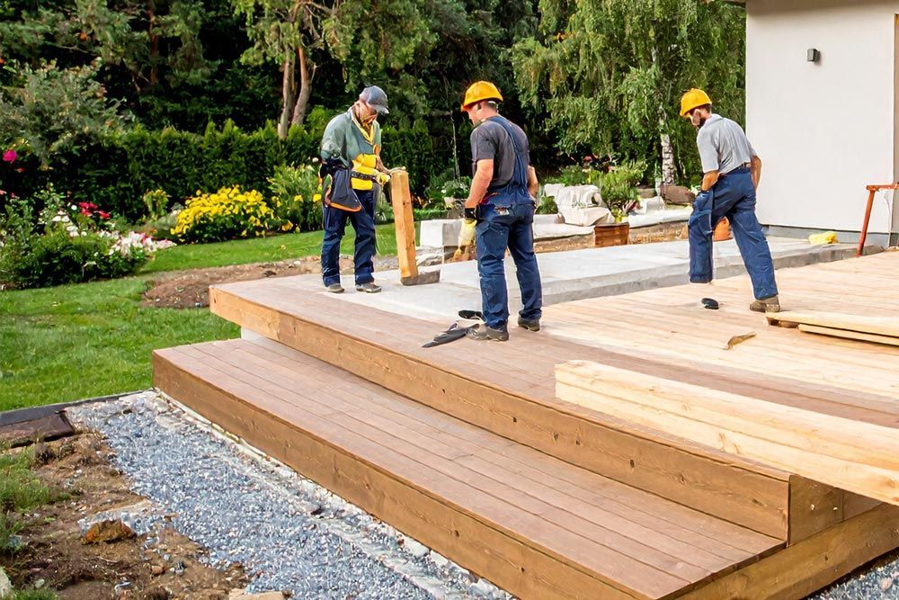 Three people building a wooden deck outdoors, wearing hard hats.