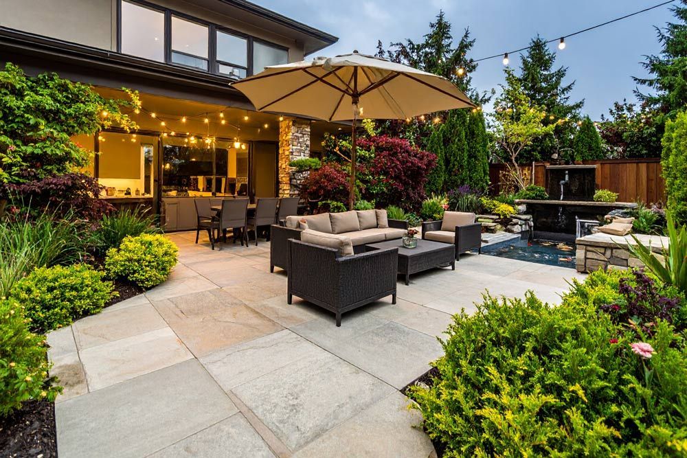 Patio with outdoor seating, umbrella, and string lights, surrounded by lush landscaping and a water feature.