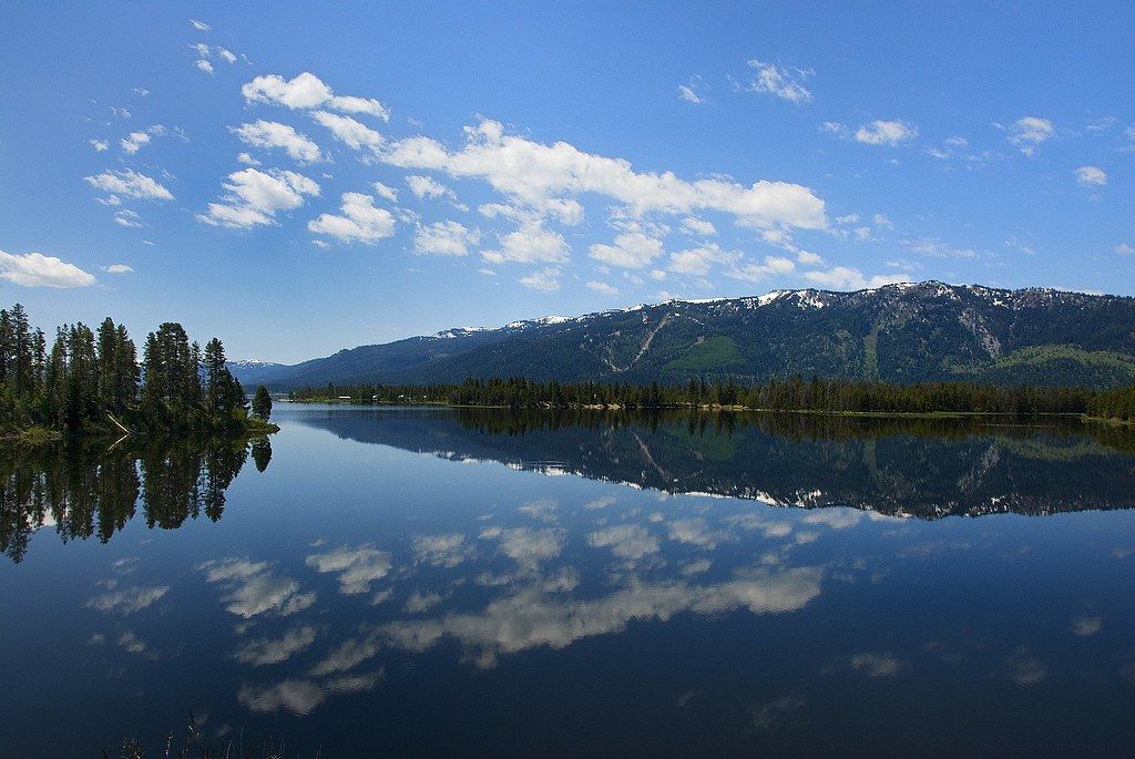 Calm lake reflects a blue sky dotted with clouds, with forested hills in the background.