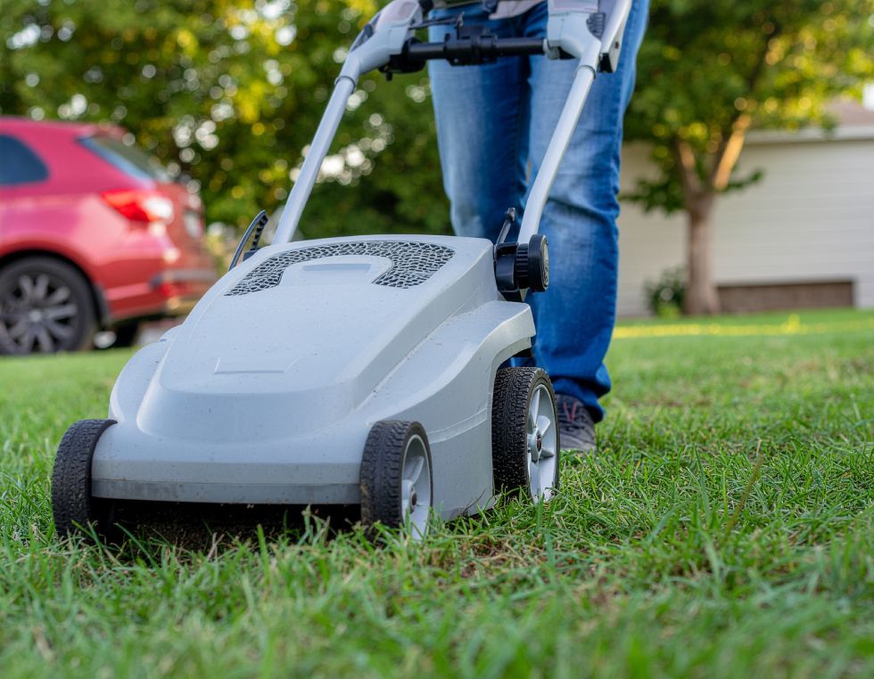 Person mowing a green lawn with a gray push mower, red car in background.