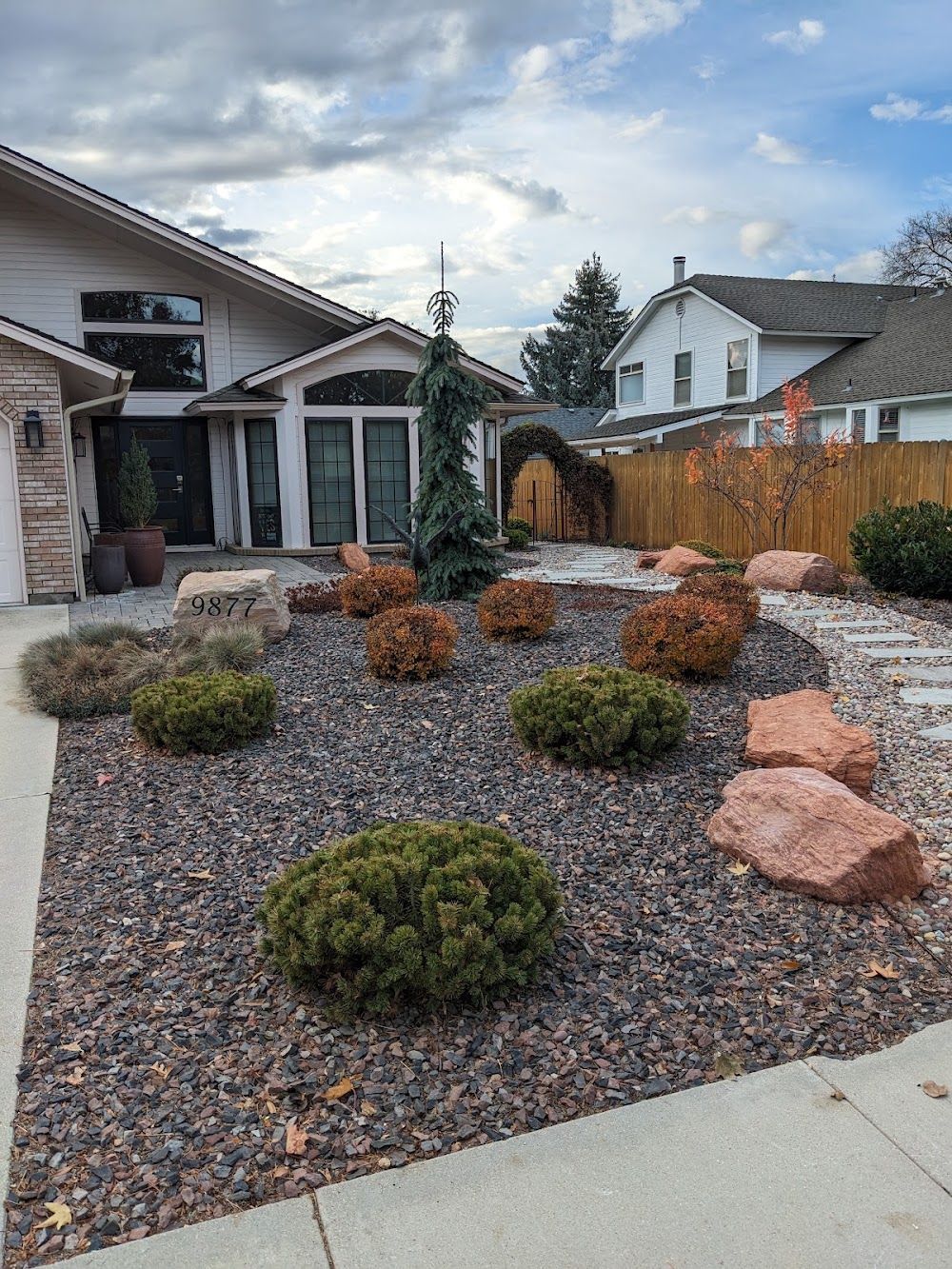 Front yard with rock landscaping, small shrubs, and a concrete path beside a house and wooden fence.