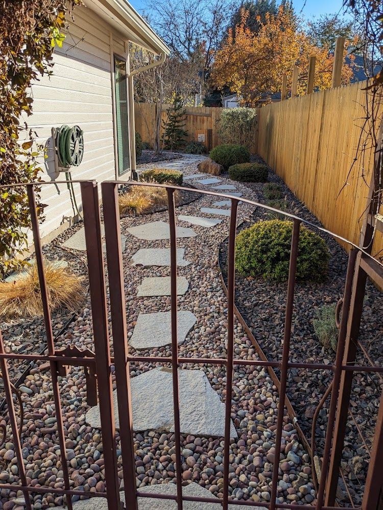 Narrow backyard gravel path with stepping stones, bordered by a wooden fence and house wall.