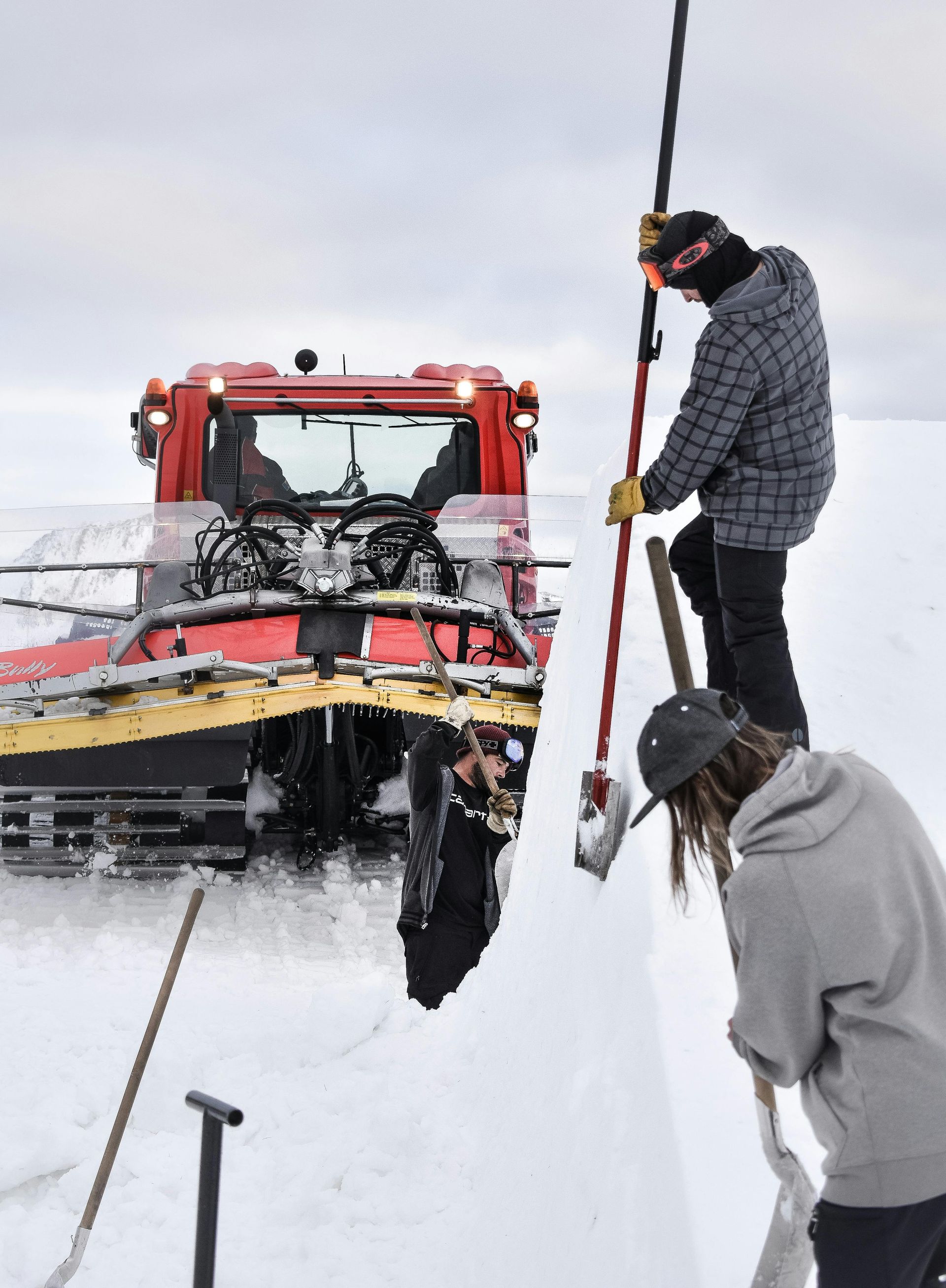 Snow groomer and people shoveling snow on a slope. Winter scene with overcast skies.