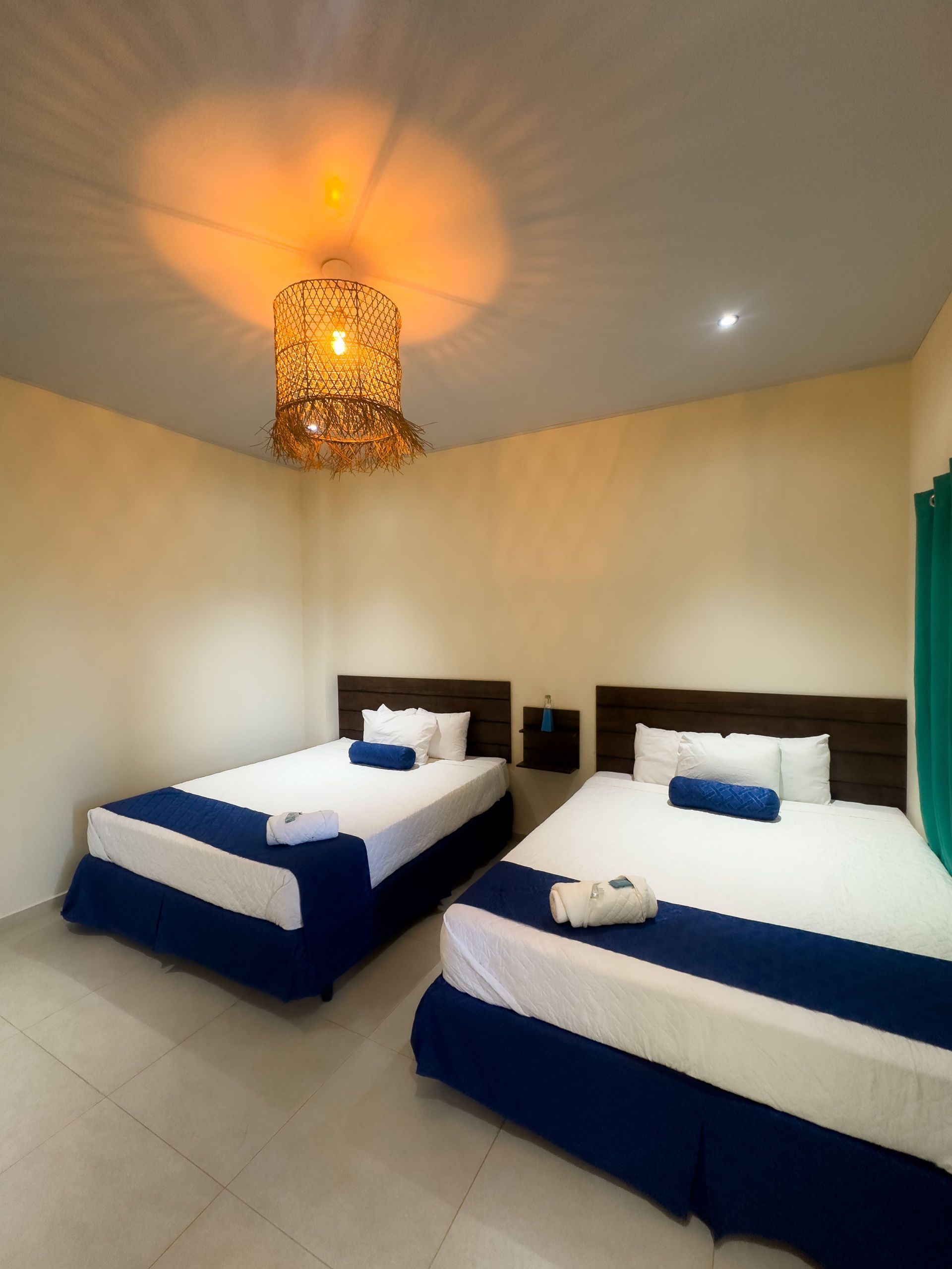 Dos camas en una habitación de hotel con ropa de cama azul y blanca, cabeceros de madera y una lámpara colgante tejida.