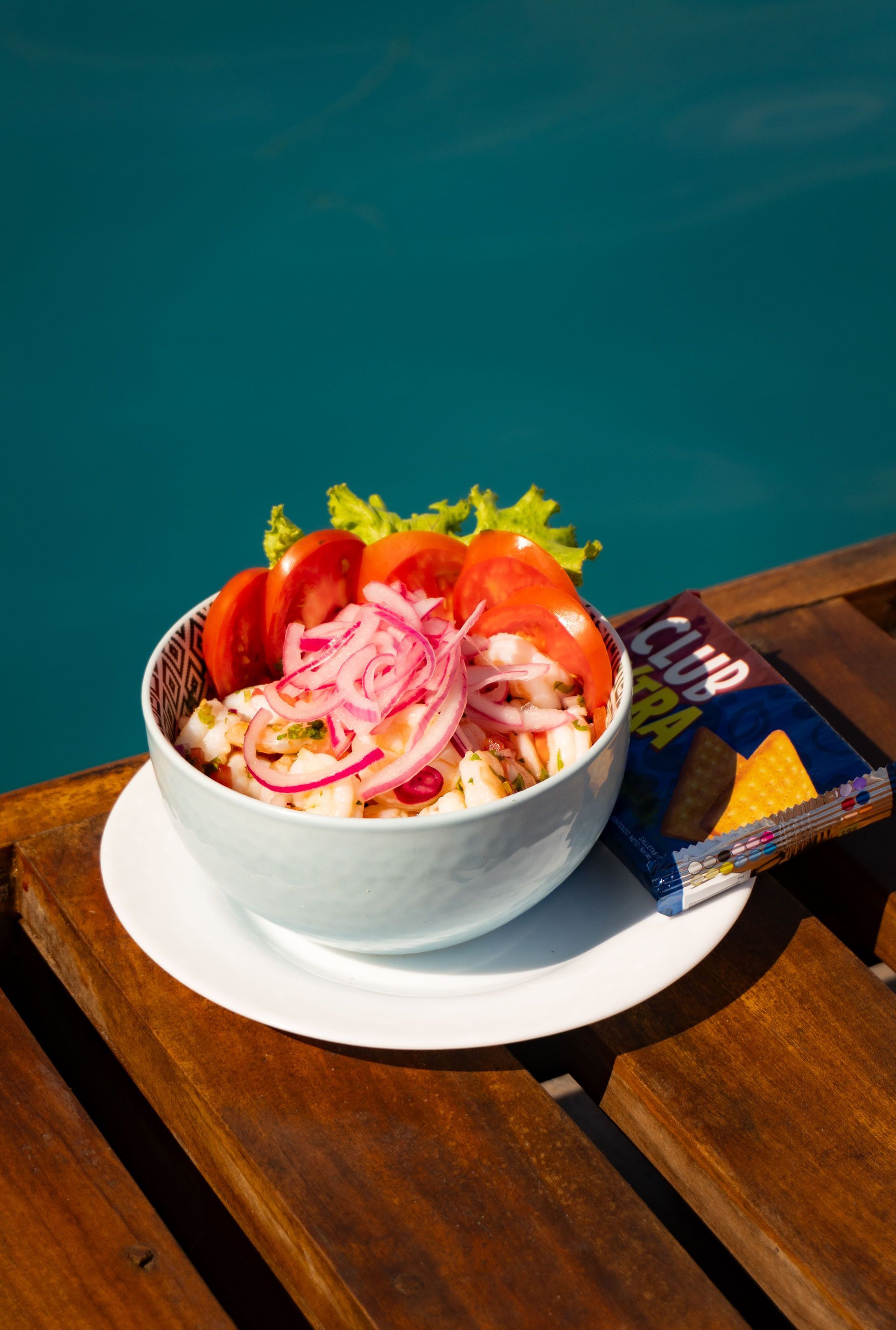 Tazón de ceviche con aderezos, papas fritas y tomate sobre una superficie de madera cerca del agua.