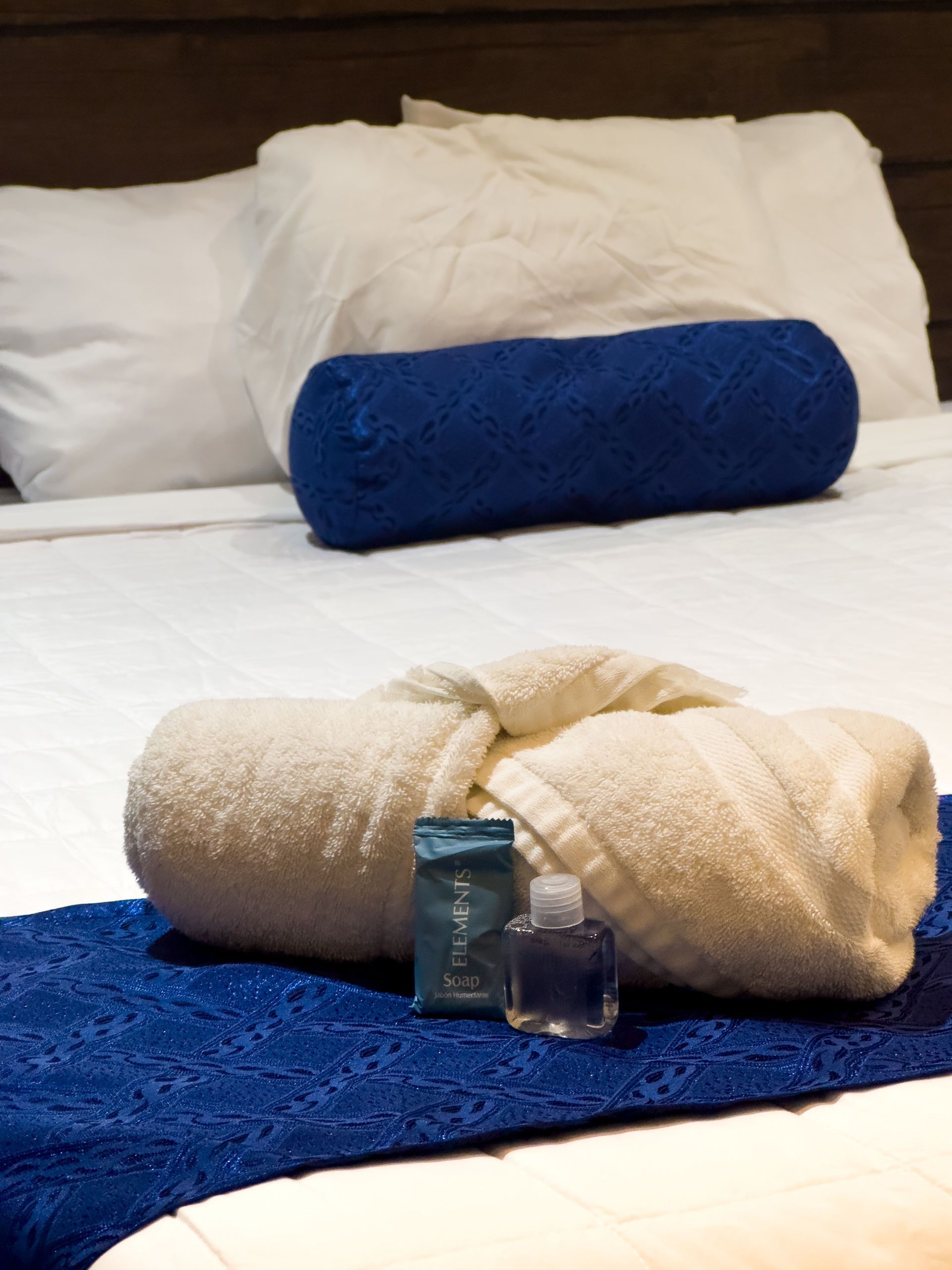 Cama blanca con camino de mesa azul, almohadas y toallas enrolladas, artículos de aseo.
