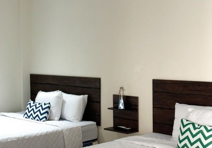 Dos camas con cabeceras de madera oscura, ropa de cama blanca y almohadas decorativas contra una pared de color crema.