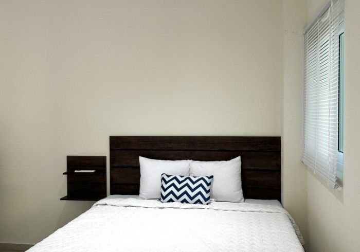 Dormitorio con cabecero de madera oscura, ropa de cama blanca, pequeña almohada azul y ventana con persianas.