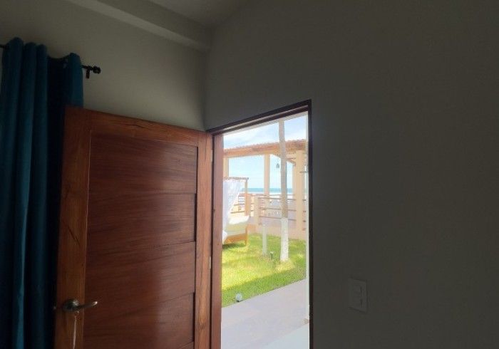 Puerta de madera parcialmente abierta, que muestra un área verde y vista al mar; cortinas verde azulado a la izquierda.