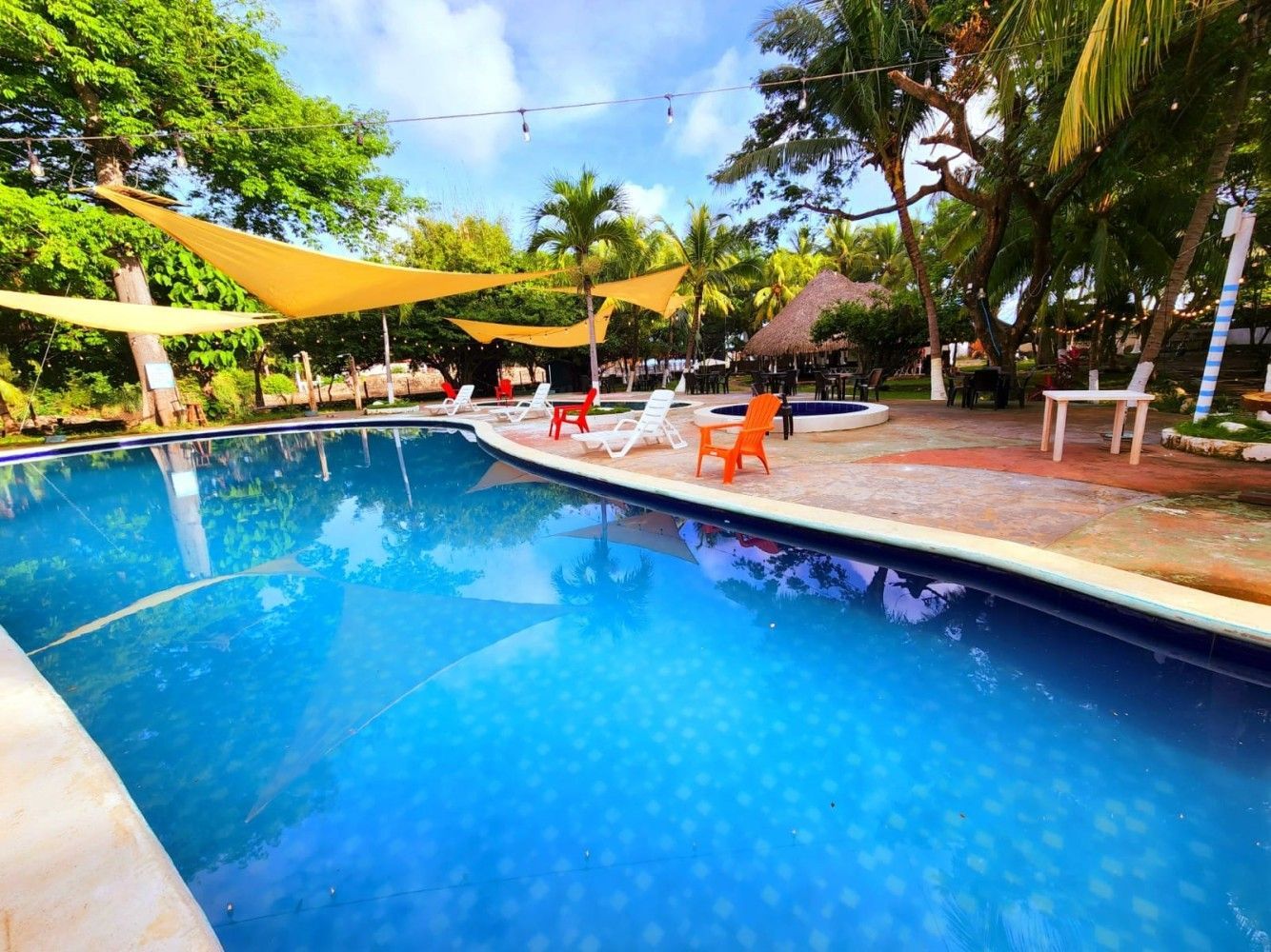 Piscina con agua azul, sillas naranjas y blancas, velas de sombra y árboles bajo un cielo azul.