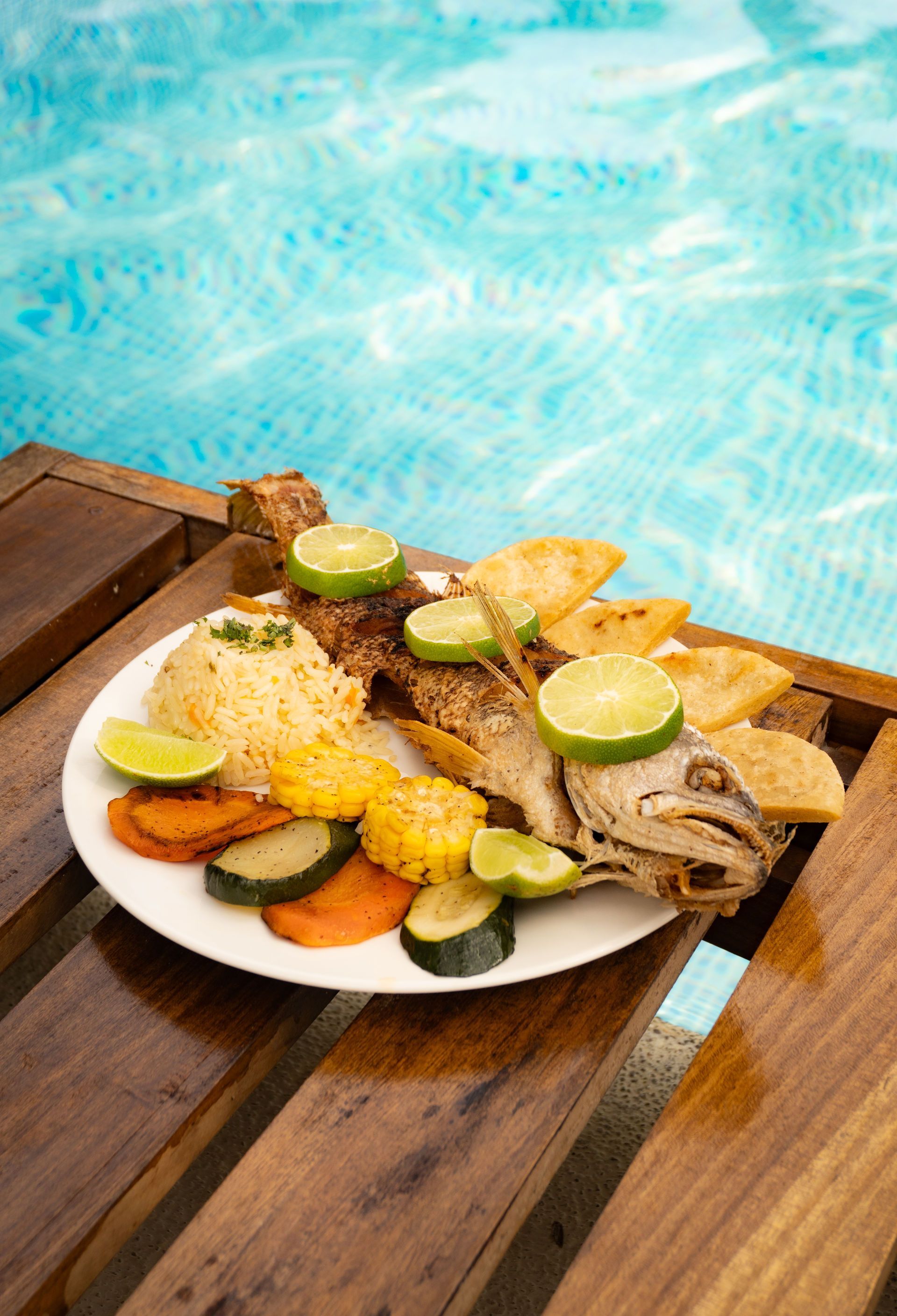 Plato de pescado frito, arroz, verduras y rodajas de lima junto a una piscina.