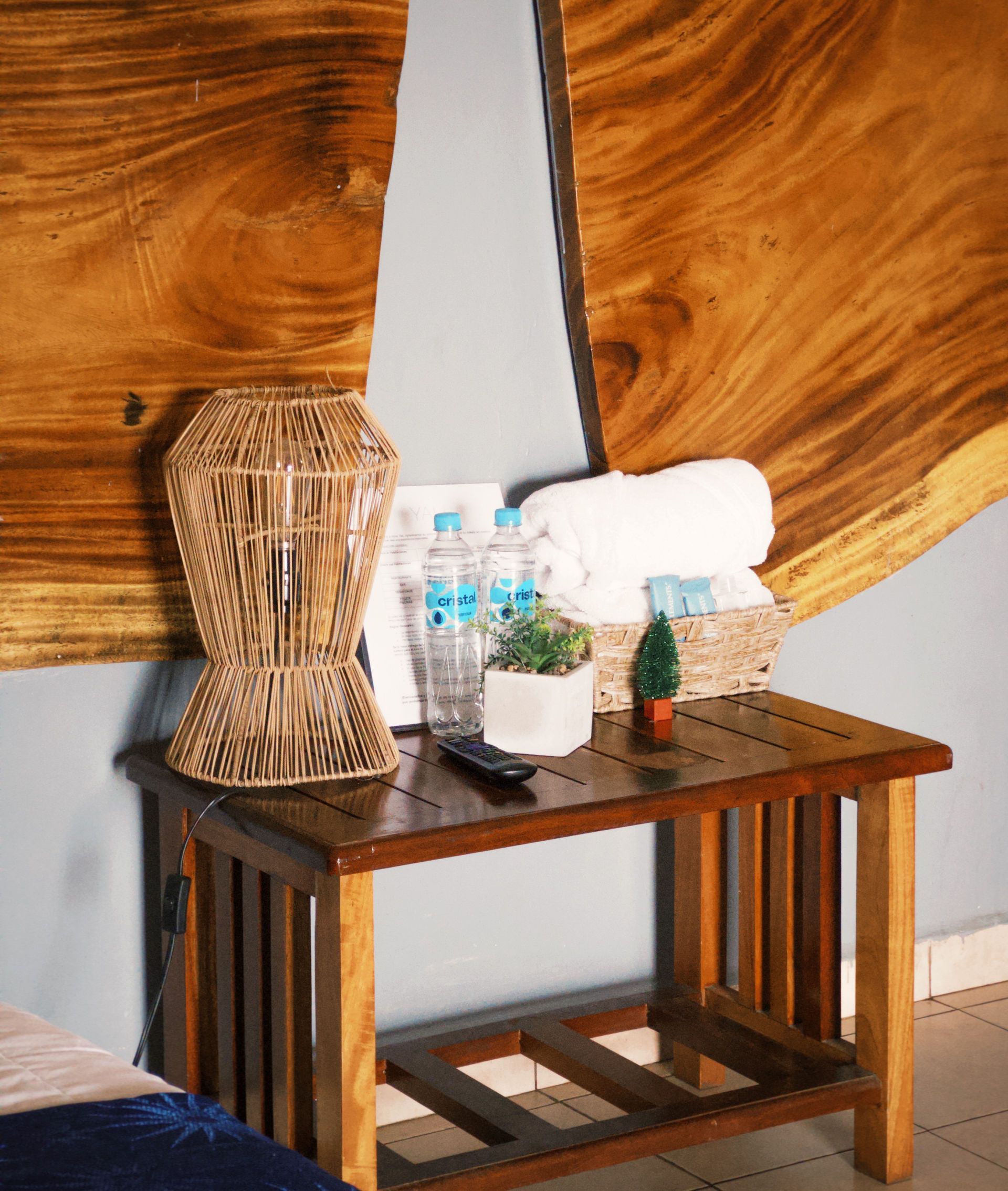 Mesita de noche con lámpara, botellas de agua, toallas y decoración, junto a una cama, contra una pared de madera.