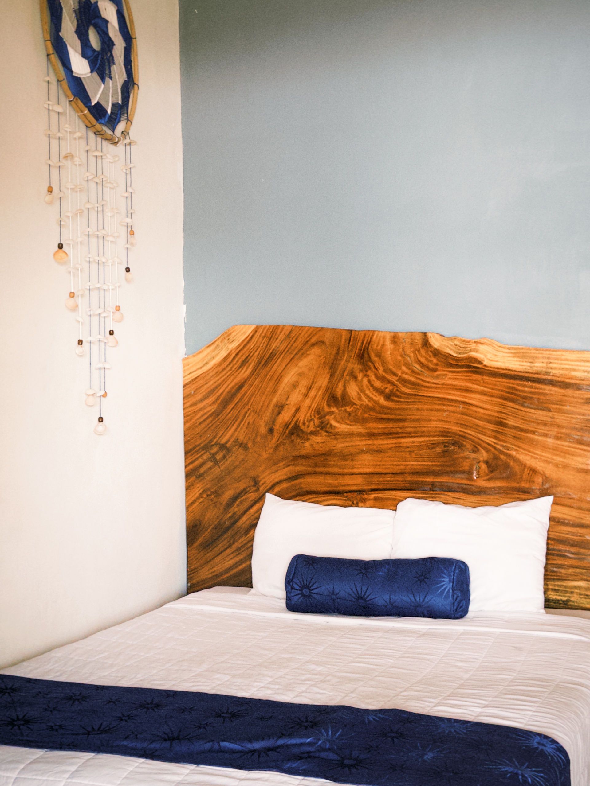 Rincón acogedor para el dormitorio. Cabecero de madera, ropa de cama blanca, detalles en azul y un atrapasueños en la pared.