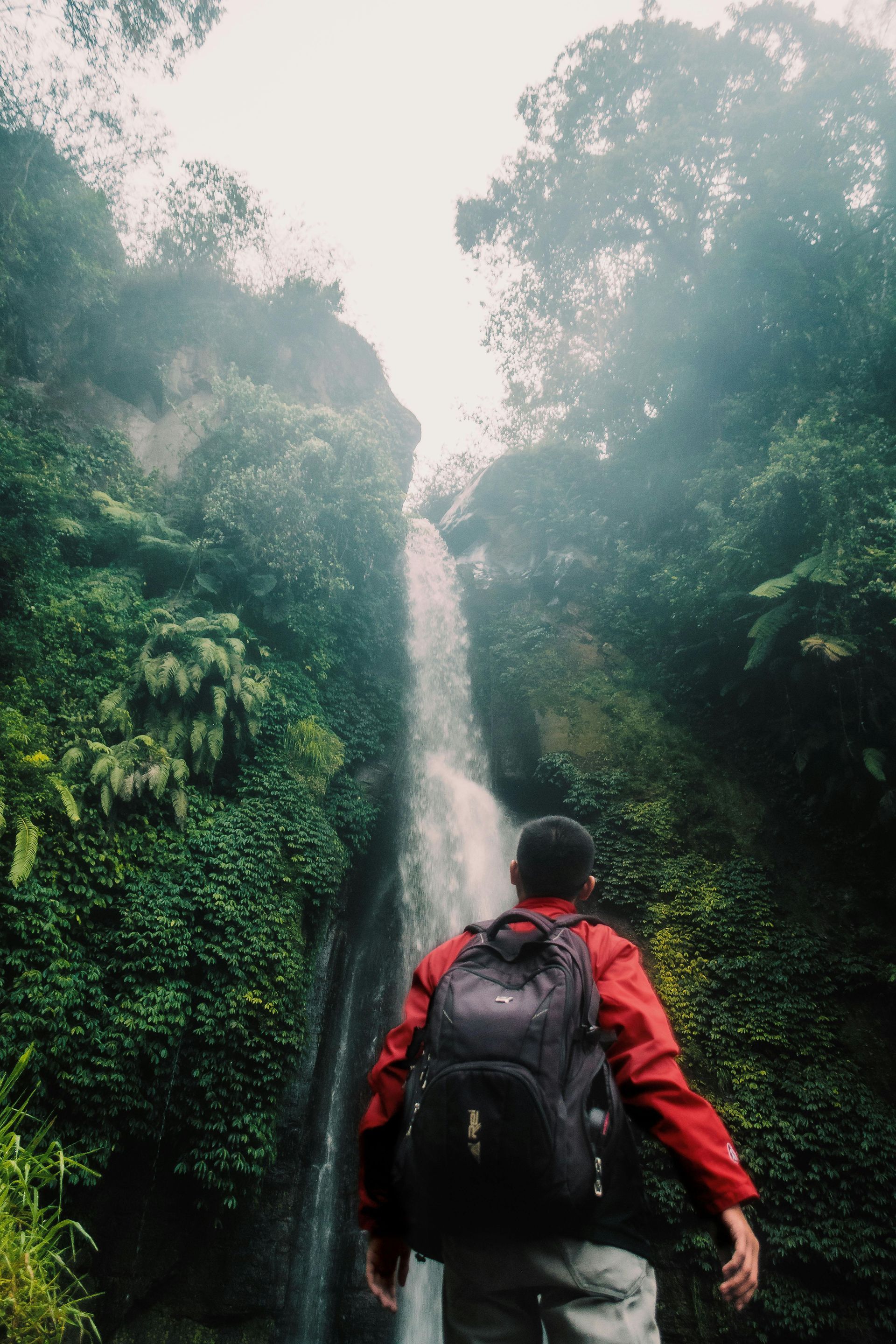Persona con mochila mirando hacia una cascada rodeada de exuberante follaje verde.