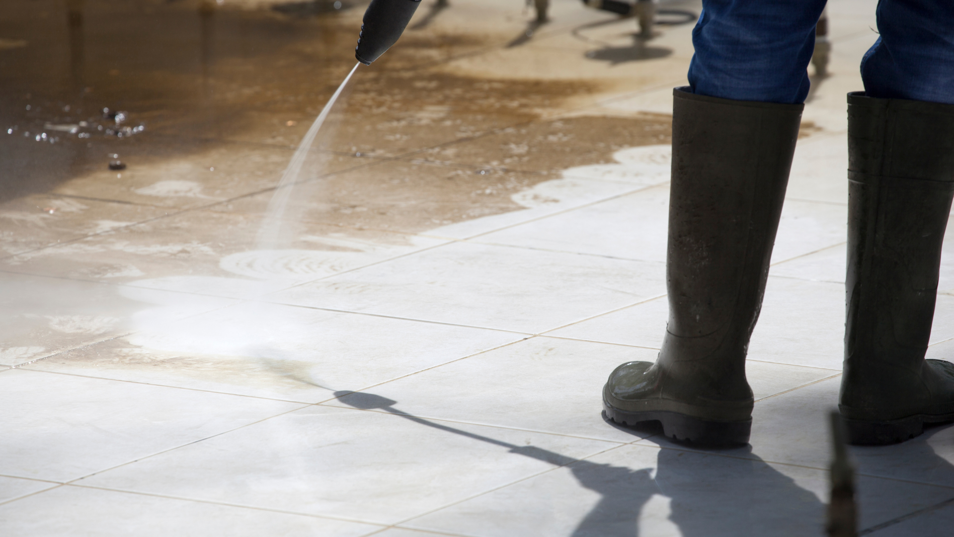 Person in tall rubber boots using a pressure washer on a dirty, tiled floor.
